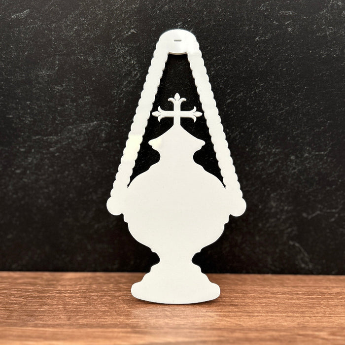 A white decorative silhouette ornament shaped like a classic oil lamp with a cross, standing on a wooden surface against a dark textured background.