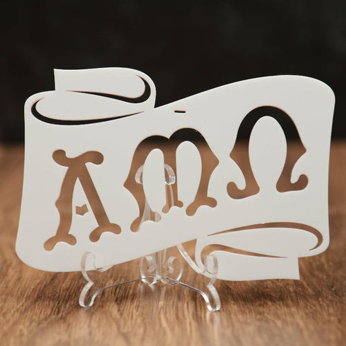 A white laser-cut acrylic scroll sign featuring the Greek letters Alpha, Mu and Omega, displayed on a clear acrylic stand over a wooden surface with a dark background.