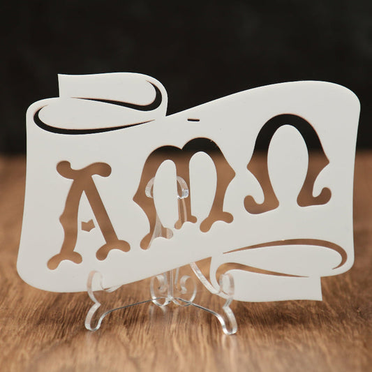 A white laser-cut acrylic scroll sign featuring the Greek letters Alpha, Mu and Omega, displayed on a clear acrylic stand over a wooden surface with a dark background.