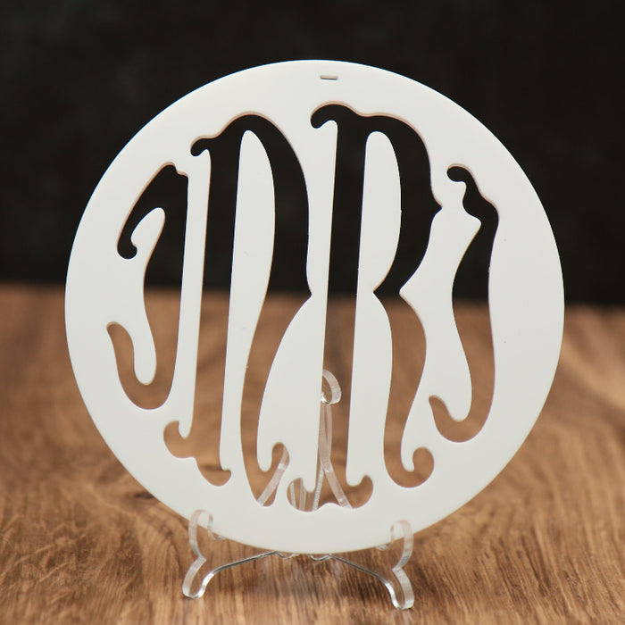 A round white monogrammed acrylic ornament with the initials 'INRI' cut out, displayed on a clear acrylic stand on a wooden surface against a dark background.