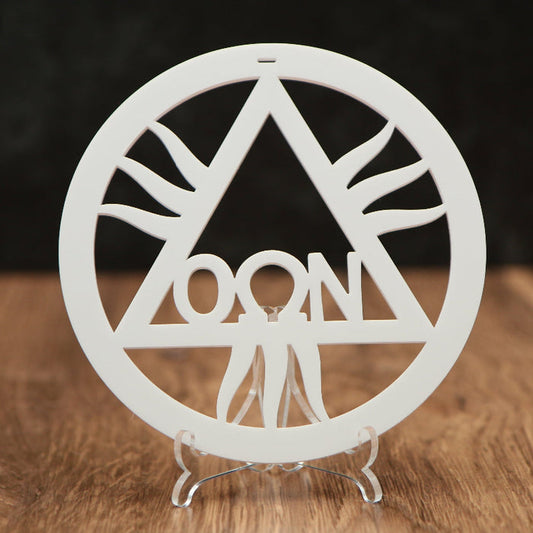 A white circular laser-cut ornament featuring a triangle with rays and the word 'OON' in the center, displayed on a clear acrylic stand on a wooden surface against a dark background.