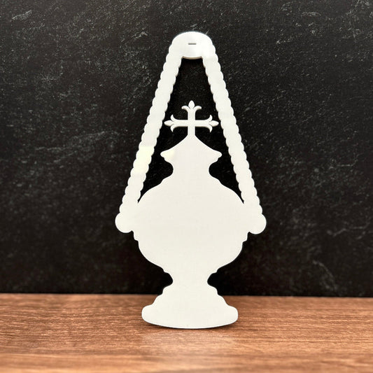 A white decorative silhouette ornament shaped like a classic oil lamp with a cross, standing on a wooden surface against a dark textured background.